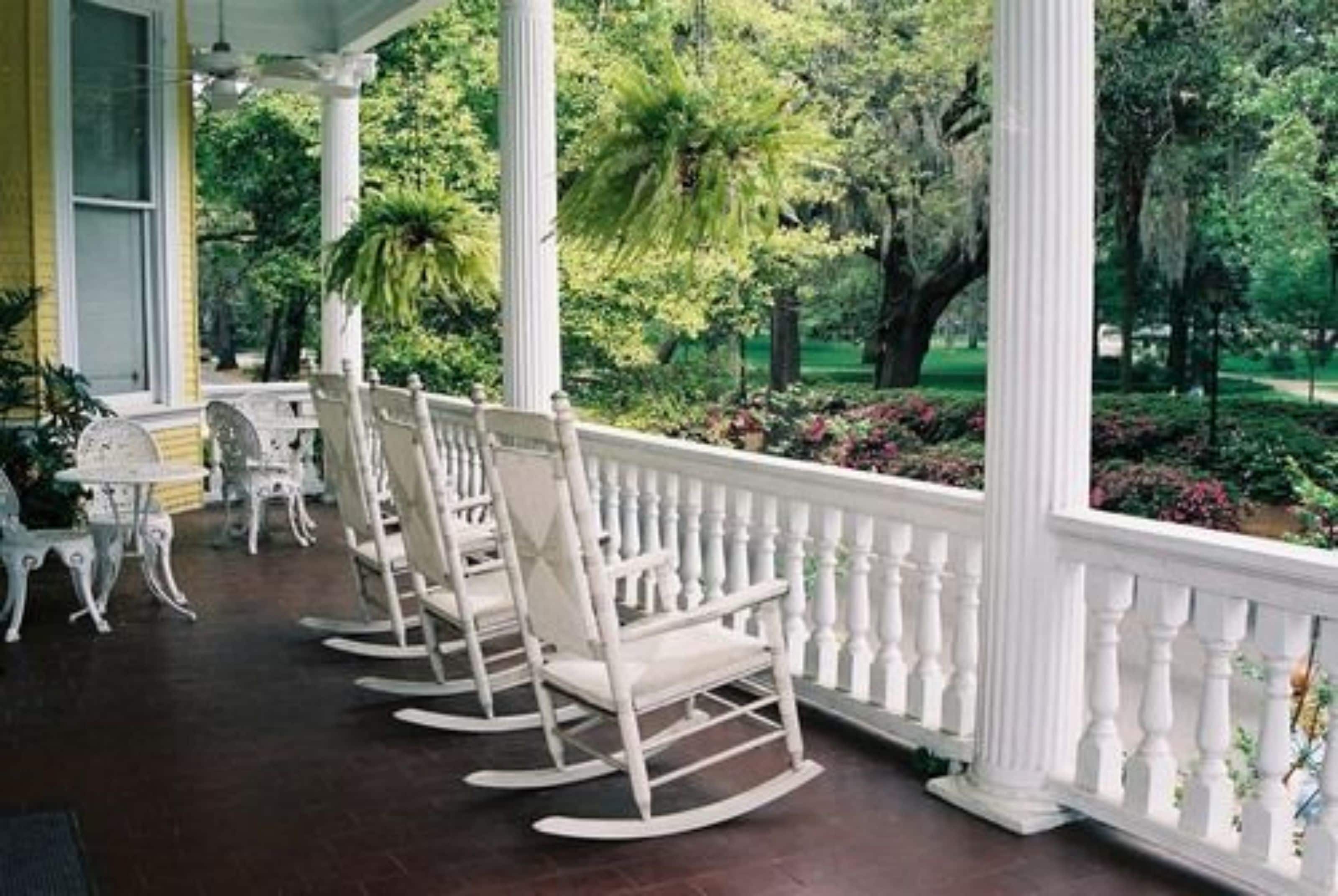 A porch with white rocking chairs and a white balustrade, surrounded by lush greenery.