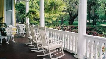 A porch with white rocking chairs and a white balustrade, surrounded by lush greenery.