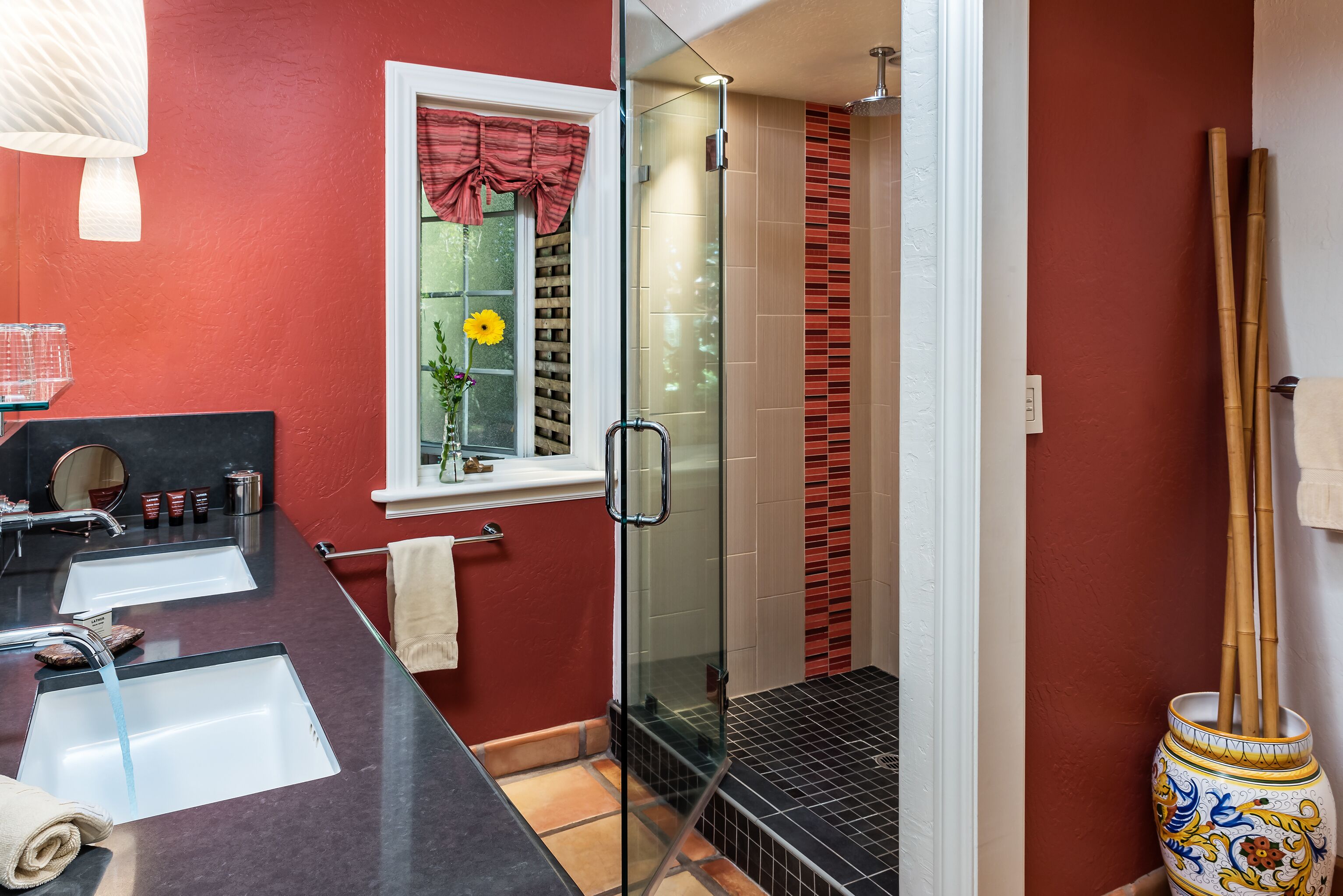 A bathroom with a glass shower enclosure, a double sink vanity, and a window with a red curtain.