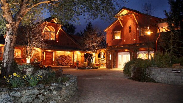 A large, two-story wooden house with a stone pathway leading to the entrance, surrounded by trees and a stone wall.
