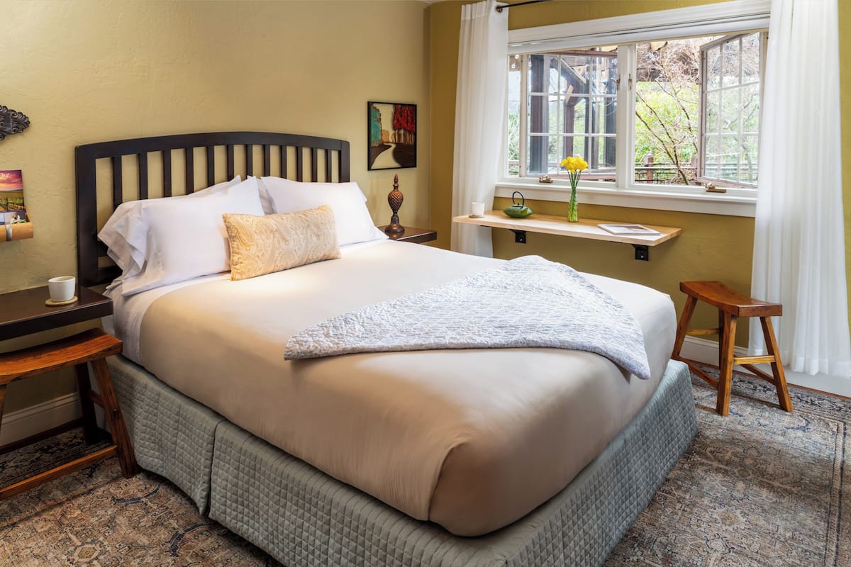 A neatly made bed with a headboard, a bedside table with a lamp, a window with a view of trees, and a small stool.