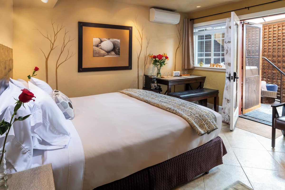 A hotel room with a bed, a desk, a vase with flowers, a framed picture on the wall, and a view of the outside.