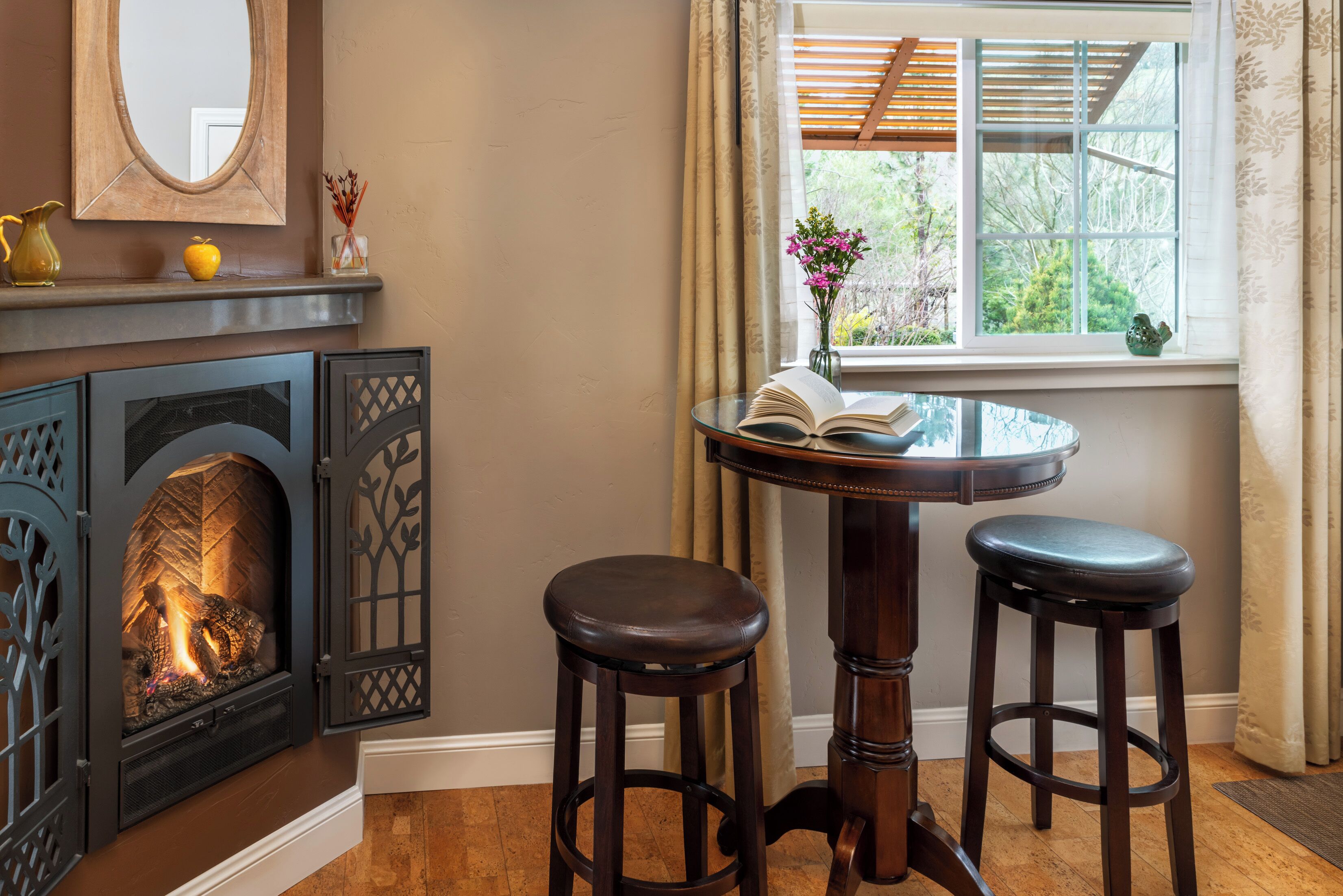 Una habitación acogedora con chimenea, una mesa redonda con dos taburetes, una ventana con vistas a la vegetación y un espejo en la pared.