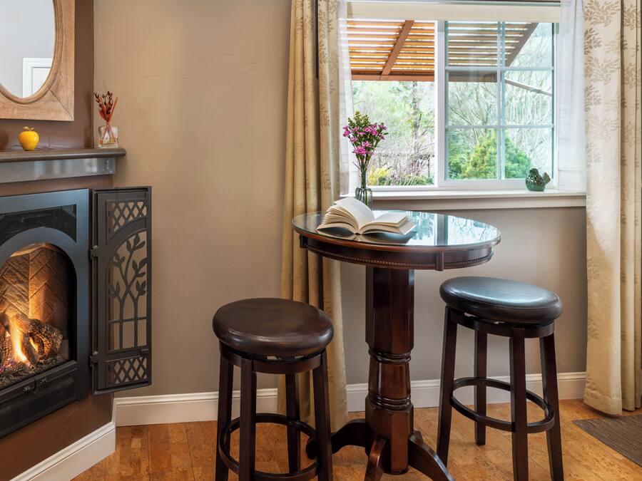 Una habitación acogedora con chimenea, una mesa redonda con dos taburetes, una ventana con vistas a la vegetación y un espejo en la pared.