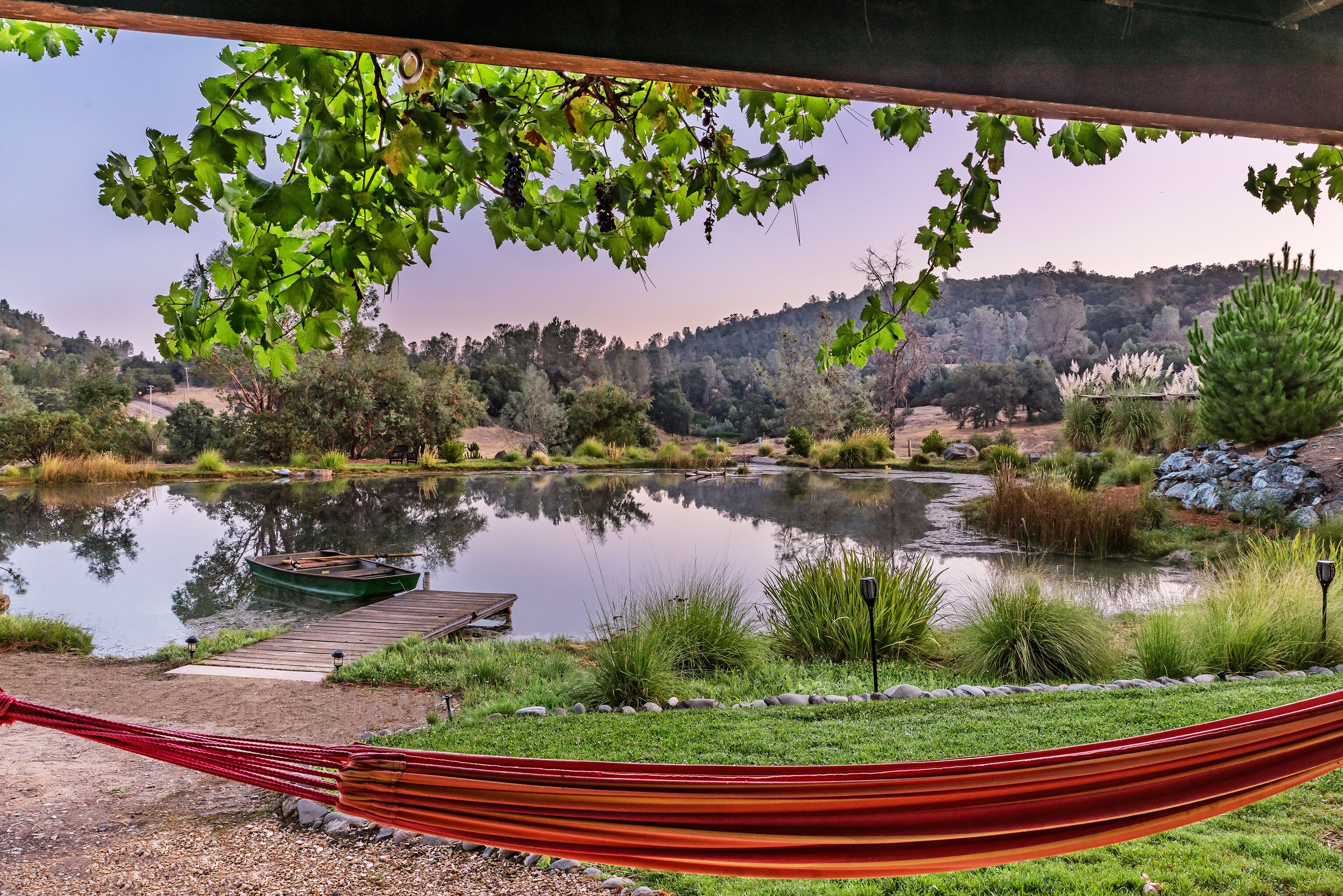 A hammock under a wooden structure with a view of a lake, dock, and surrounding greenery.