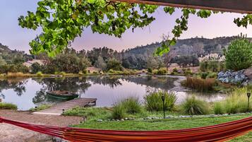 A hammock under a wooden structure with a view of a lake, dock, and surrounding greenery.