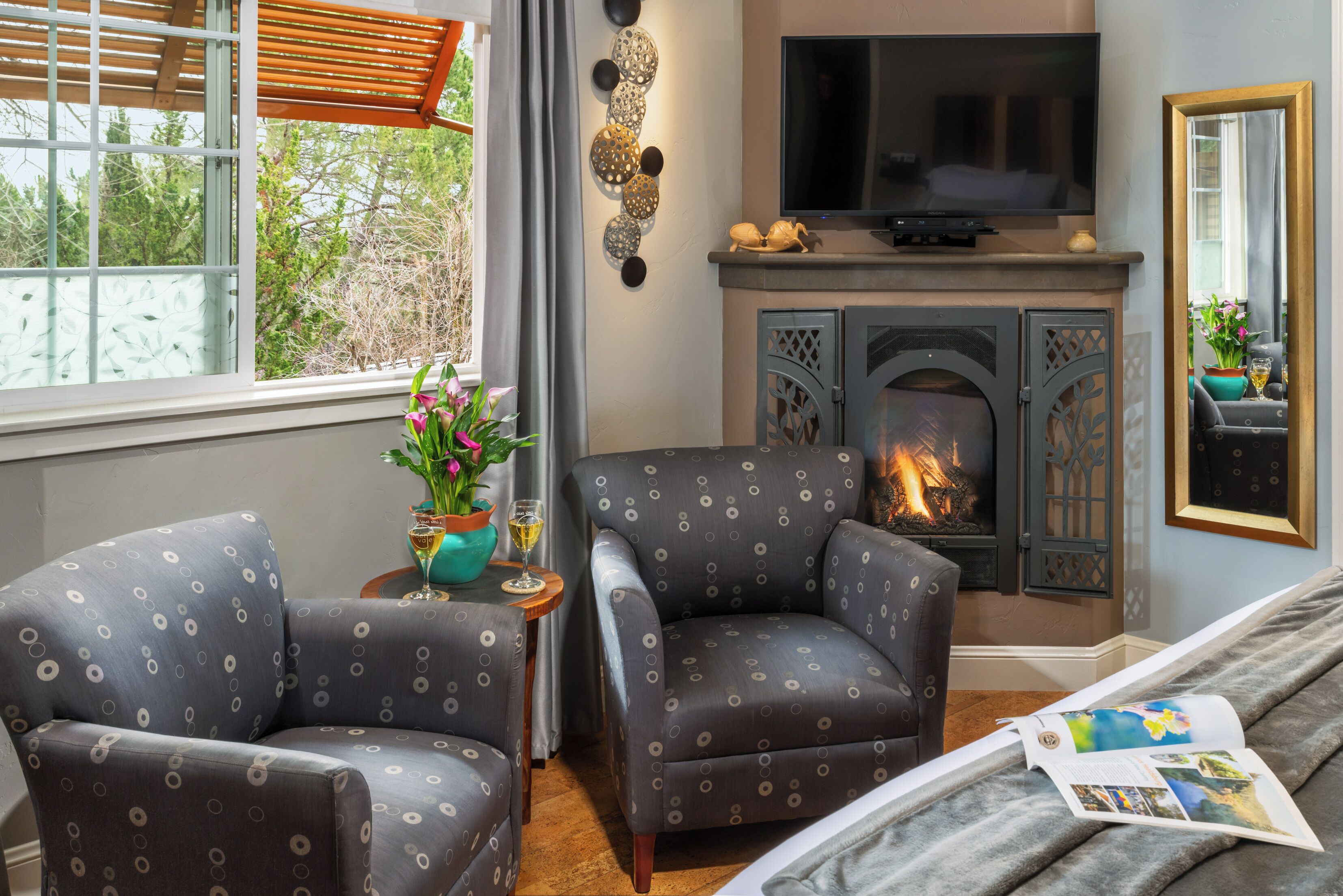 A cozy living room with a fireplace, two patterned armchairs, a TV, and a window with a view of greenery.