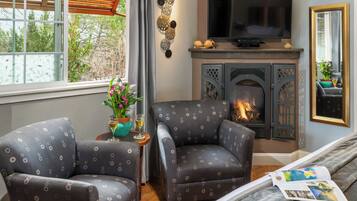 A cozy living room with a fireplace, two patterned armchairs, a TV, and a window with a view of greenery.