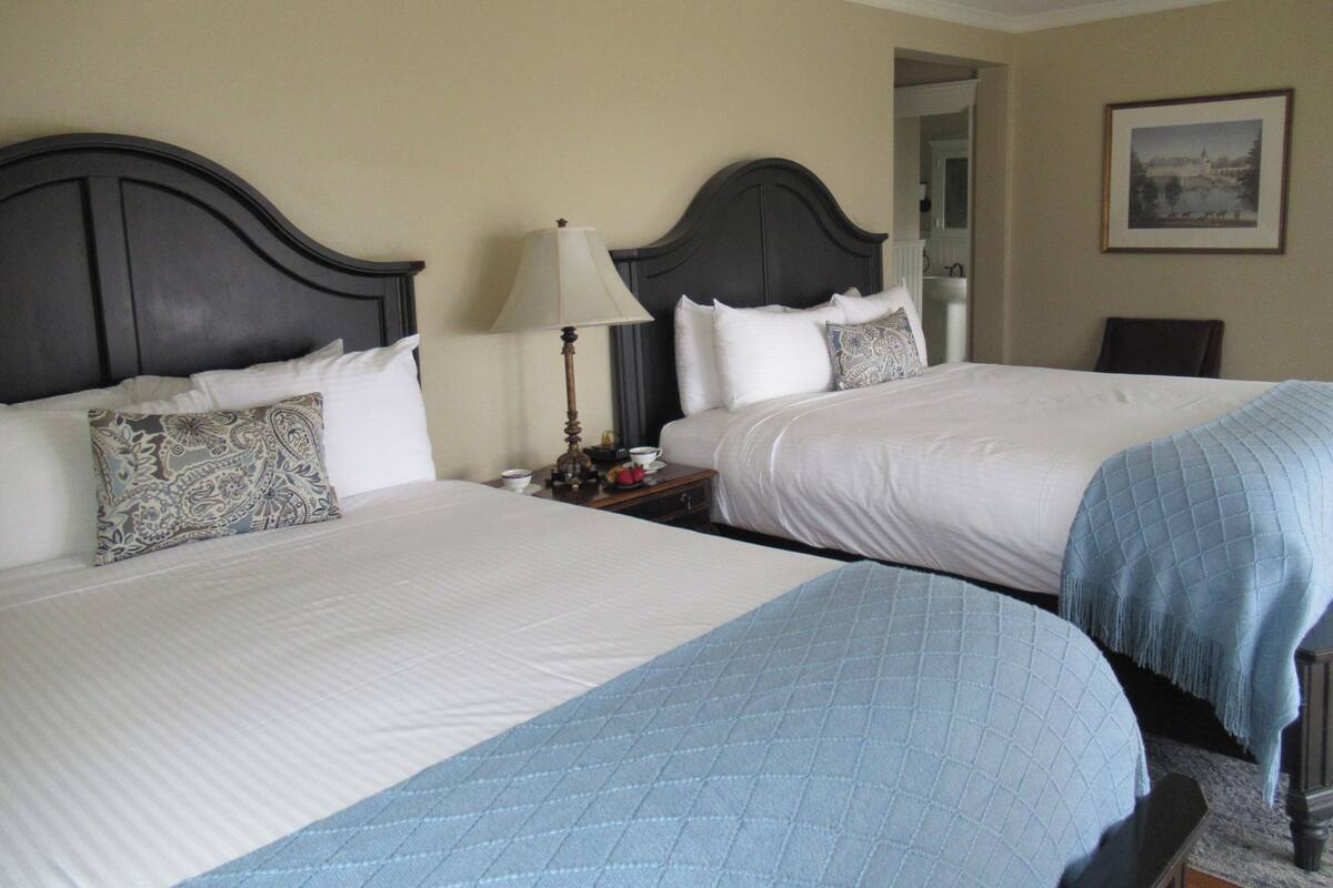 Quarto de hotel com duas camas, uma mesinha de cabeceira com abajur e um quadro na parede.