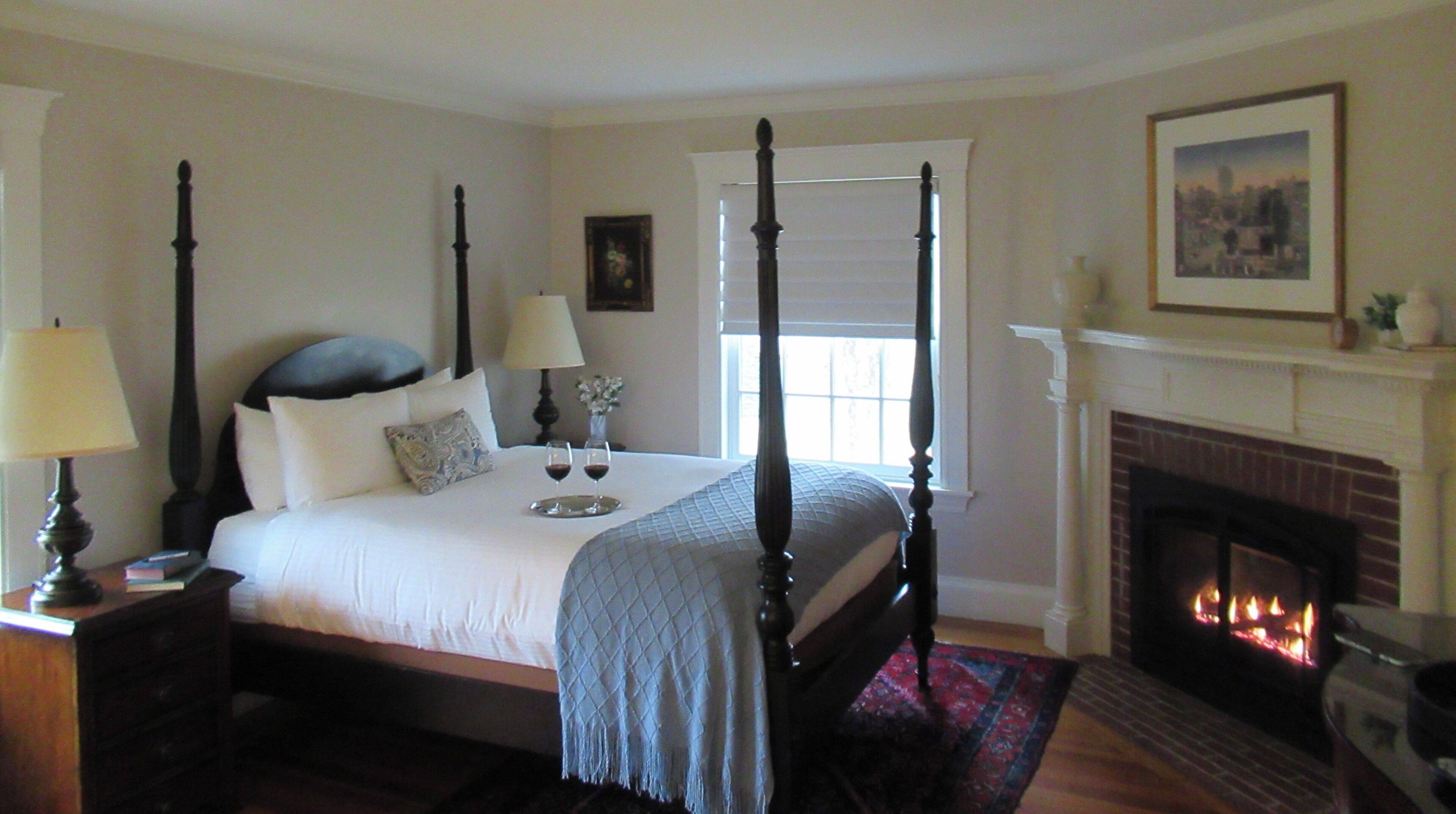 A four-poster bed with a white bedspread, a bedside table with a lamp, a vase with flowers, and a fireplace with a fire.