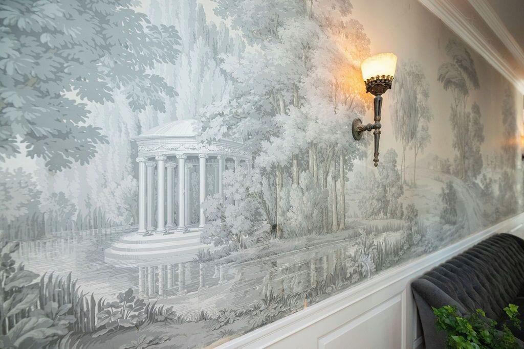 A mural with a classical building and trees, a wall-mounted light fixture, and a cushioned bench.