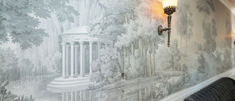 A mural with a classical building and trees, a wall-mounted light fixture, and a cushioned bench.