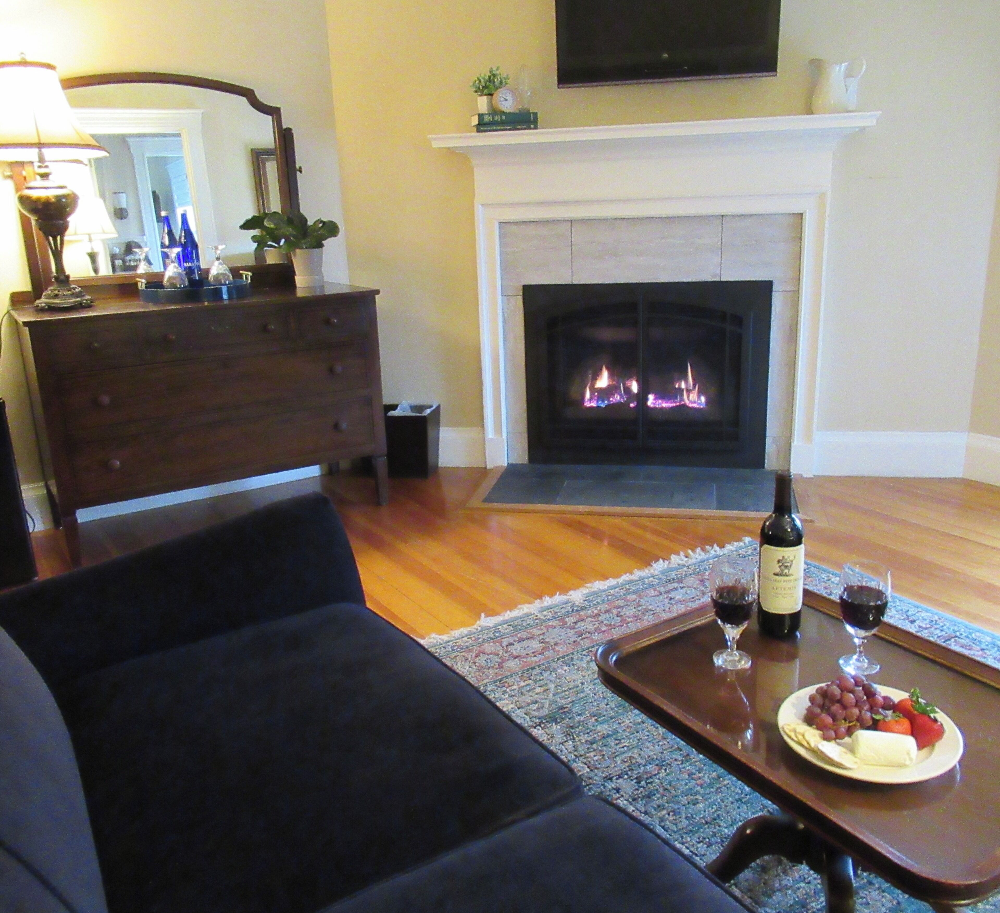 A living room with a fireplace, a sofa, a coffee table with wine glasses and grapes, and a television on the wall.