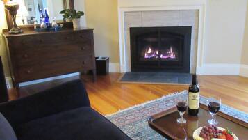 A living room with a fireplace, a sofa, a coffee table with wine glasses and grapes, and a television on the wall.