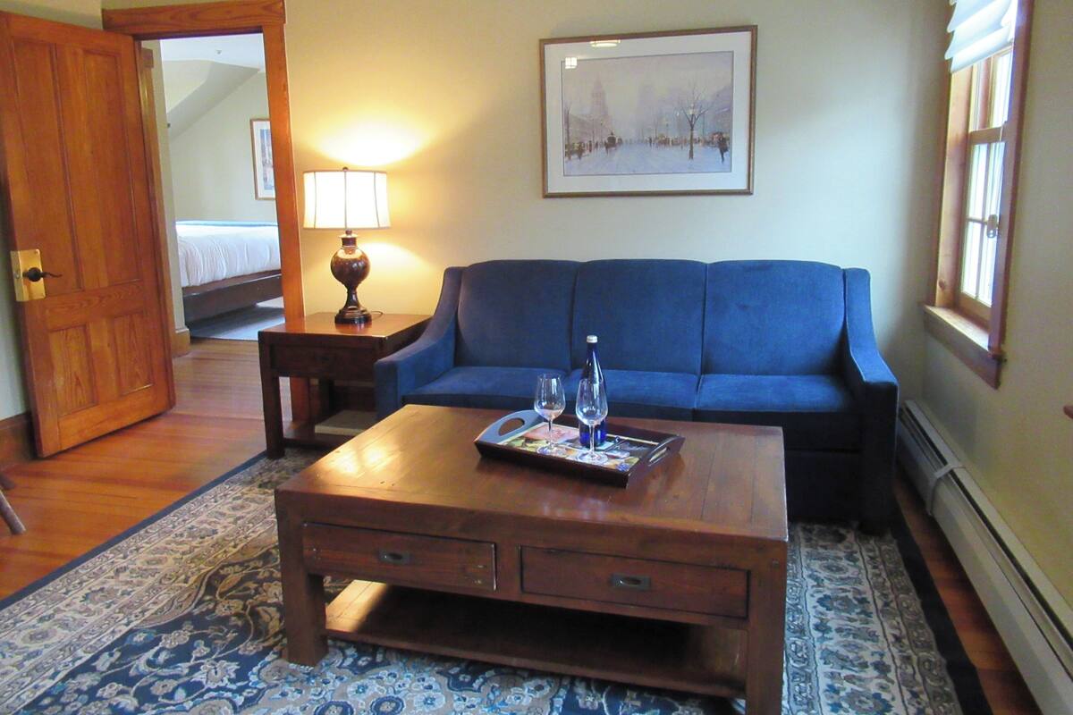 Uma sala de estar com um sofá azul, uma mesa de centro de madeira com um tabuleiro, um abajur e um quadro na parede.
