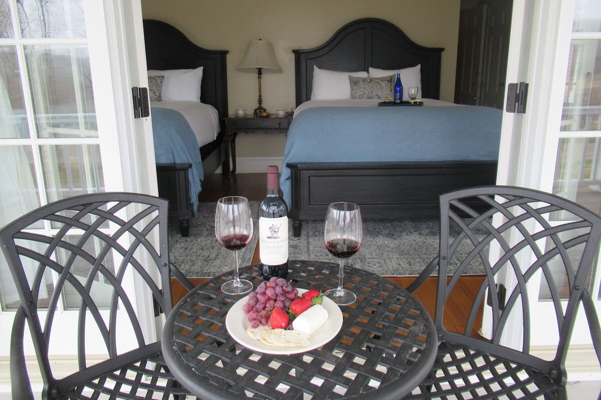 Quarto de hotel com duas camas, uma mesa de jantar com uma travessa de fruta e copos de vinho, e vista para o exterior.