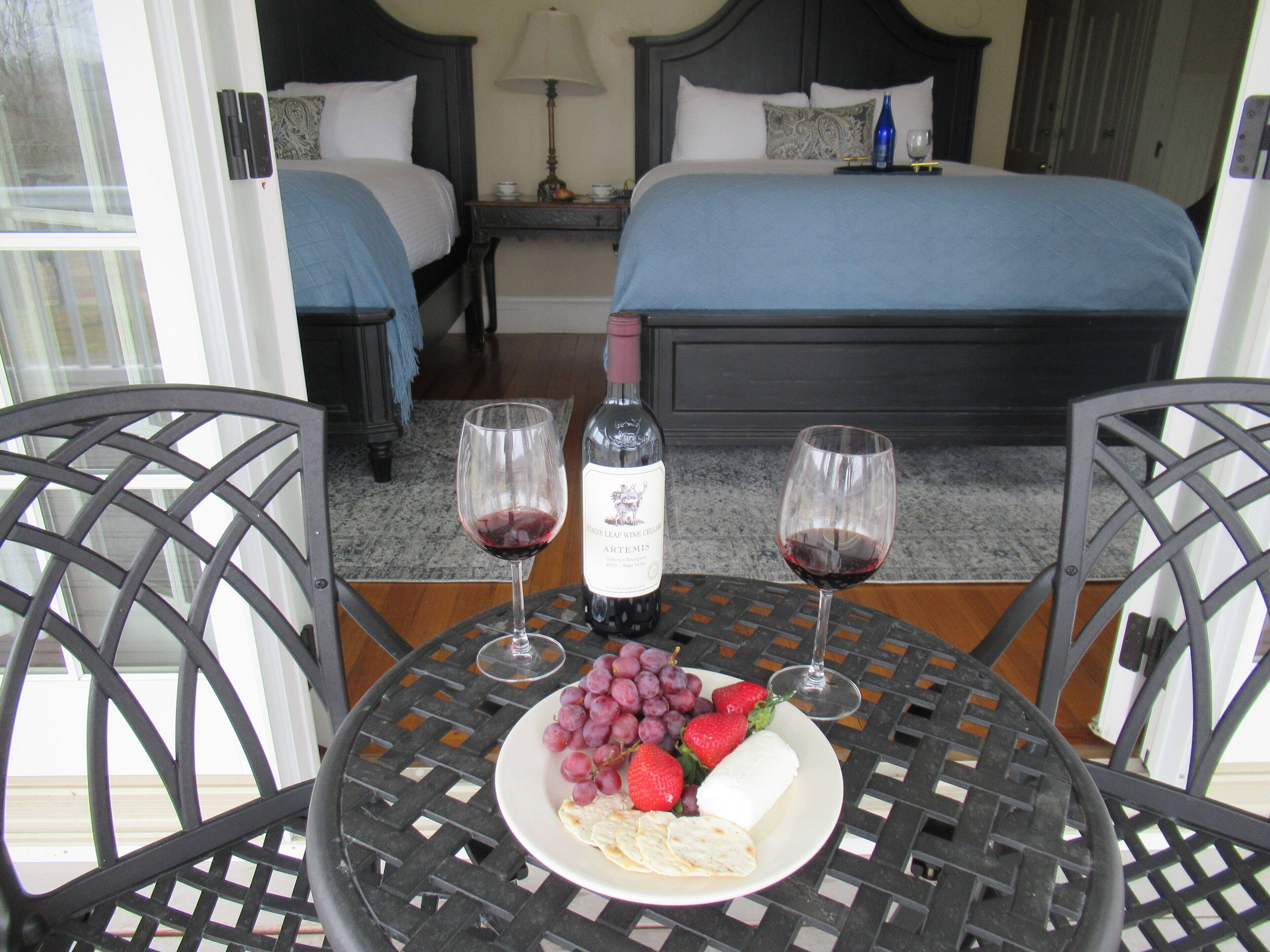 Una camera d'albergo con un letto, due comodini, un tavolino da notte e un balcone con un tavolo, una bottiglia di vino, due bicchieri e un piatto di uva e fragole.