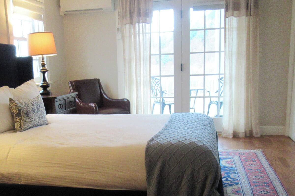 Quarto de hotel com uma cama grande, uma cadeira, uma mesa e vista para o exterior através de uma janela.