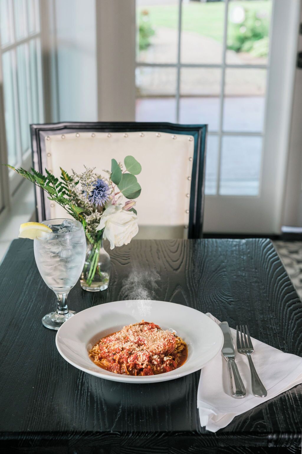 A dining table with a plate of pasta, a glass of water with a lemon slice, and a vase of flowers.