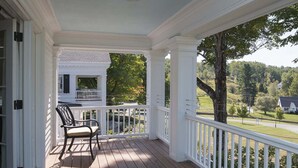 Eine überdachte Veranda mit Holzboden und weißen Geländern, ein schwarzer Sessel und im Hintergrund ein weißes Haus sowie Bäume.