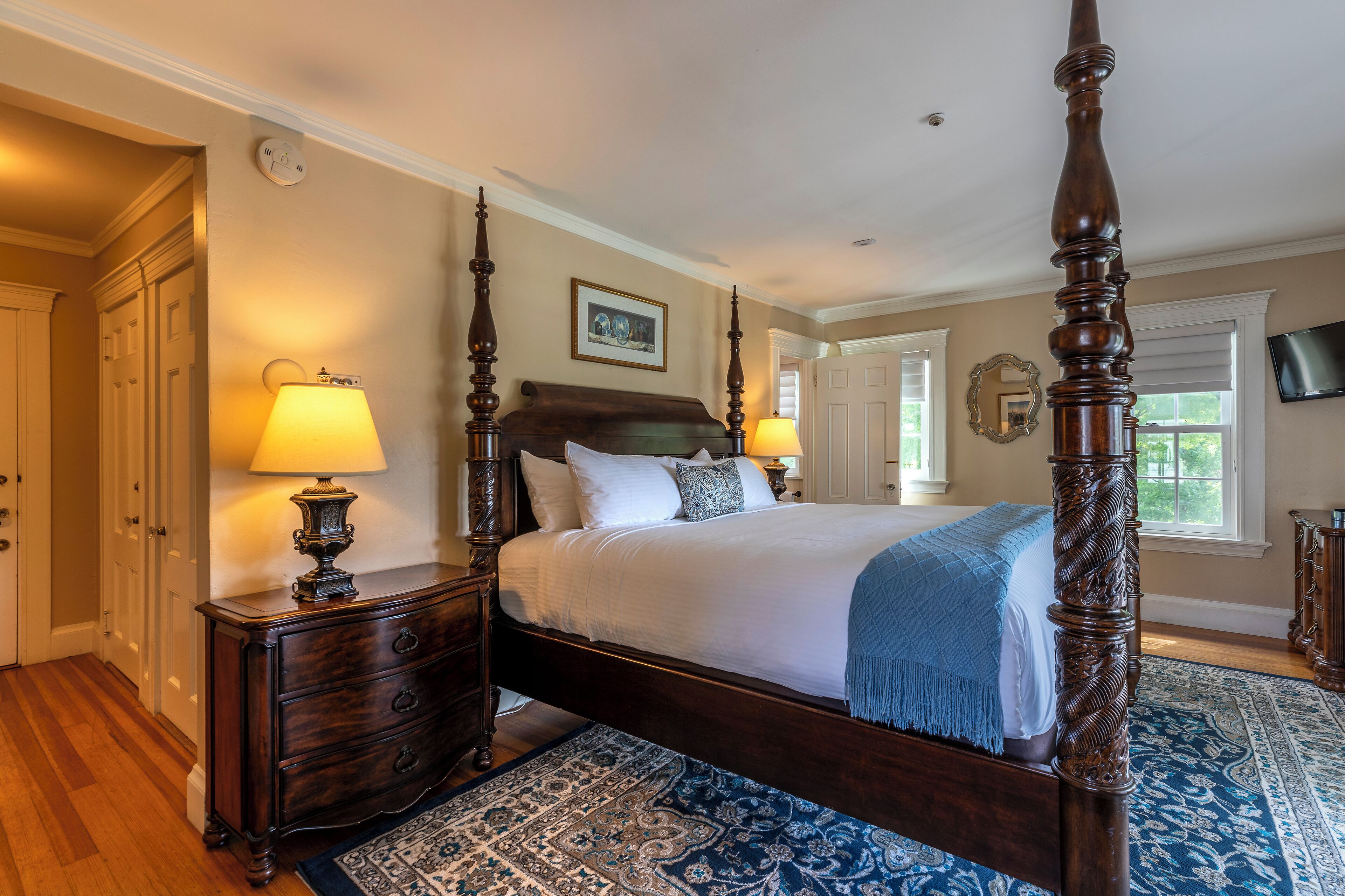 A bedroom with a four-poster bed, two bedside lamps, a wooden dresser, and a patterned rug.