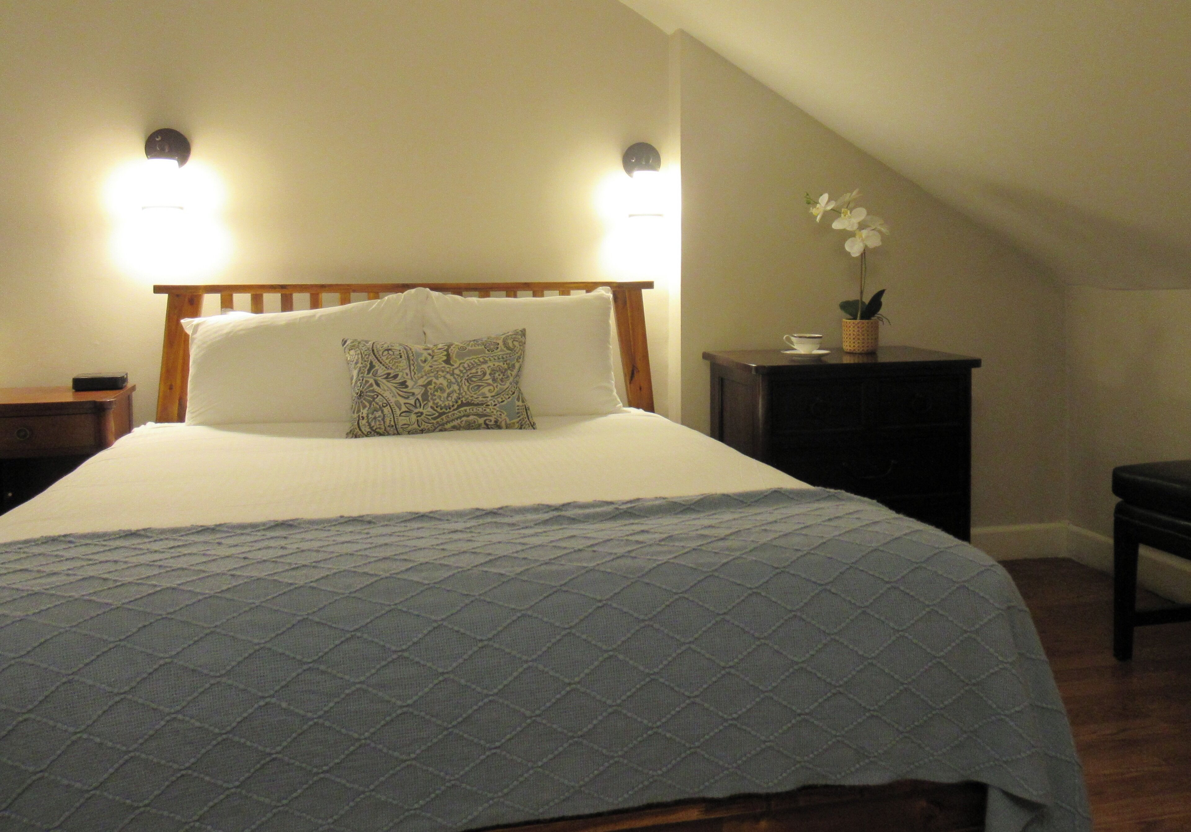 A neatly made bed with a quilted cover, two bedside lamps, a small nightstand, and a bedside table with a flower vase.
