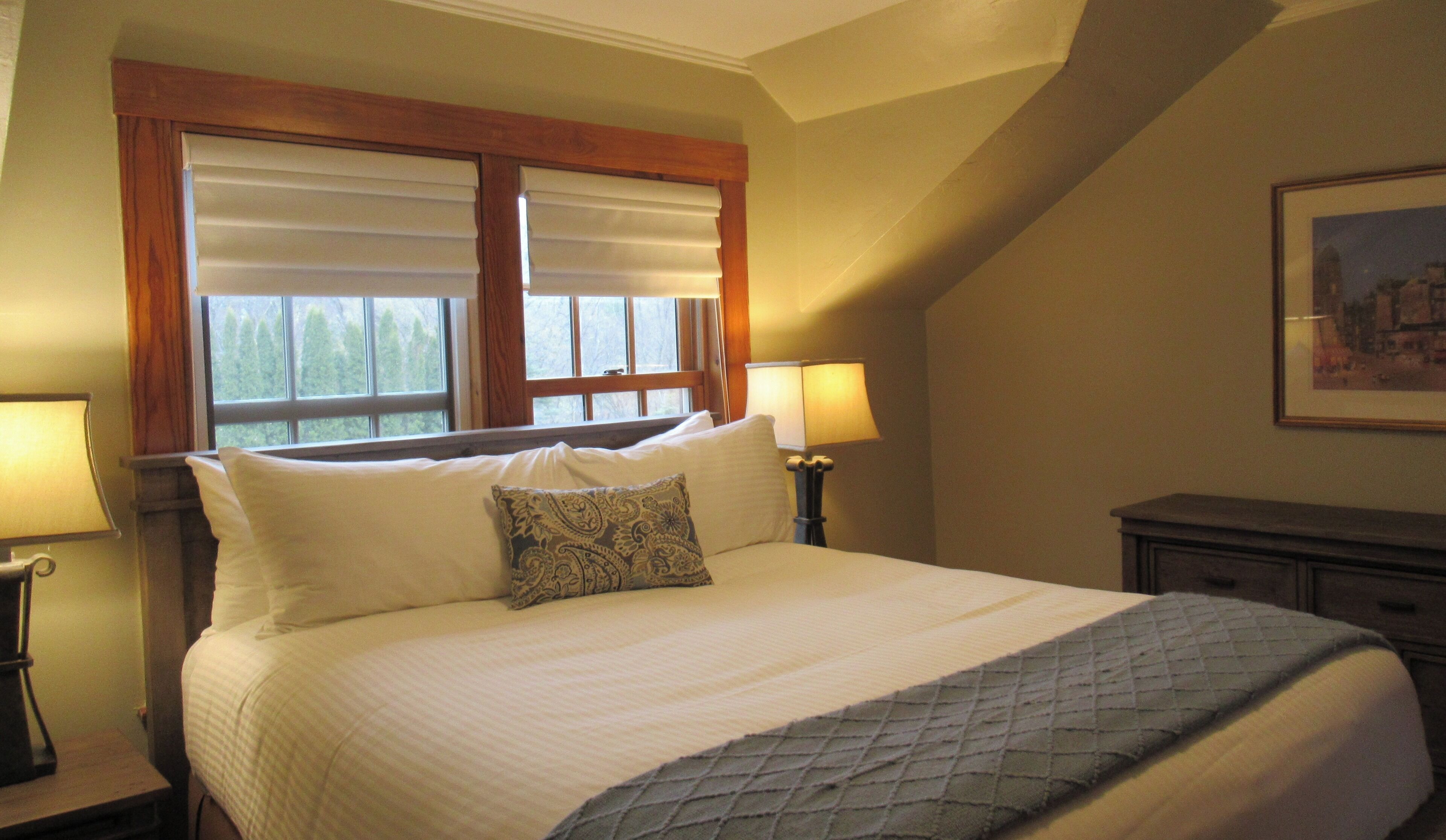 A bedroom with a large bed, two bedside lamps, a window with blinds, and a framed picture on the wall.