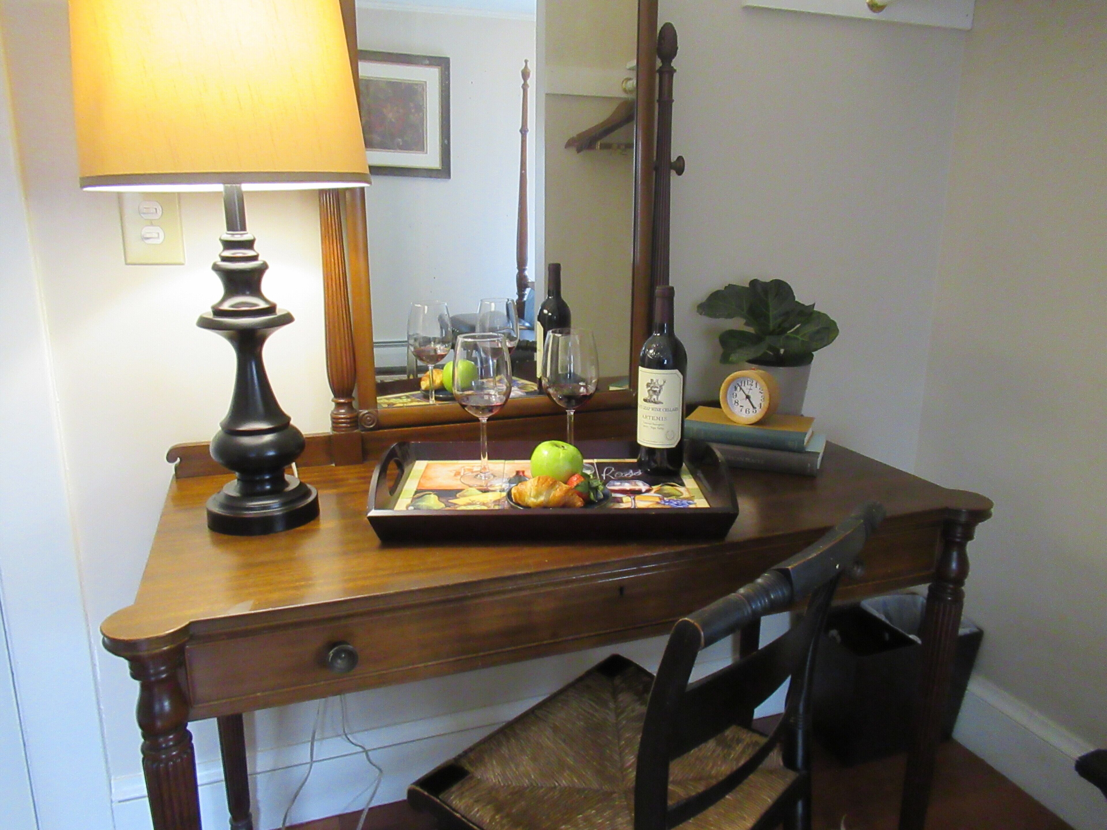 Un escritorio de madera con una lámpara, una bandeja con comida y vino, un reloj y un espejo.