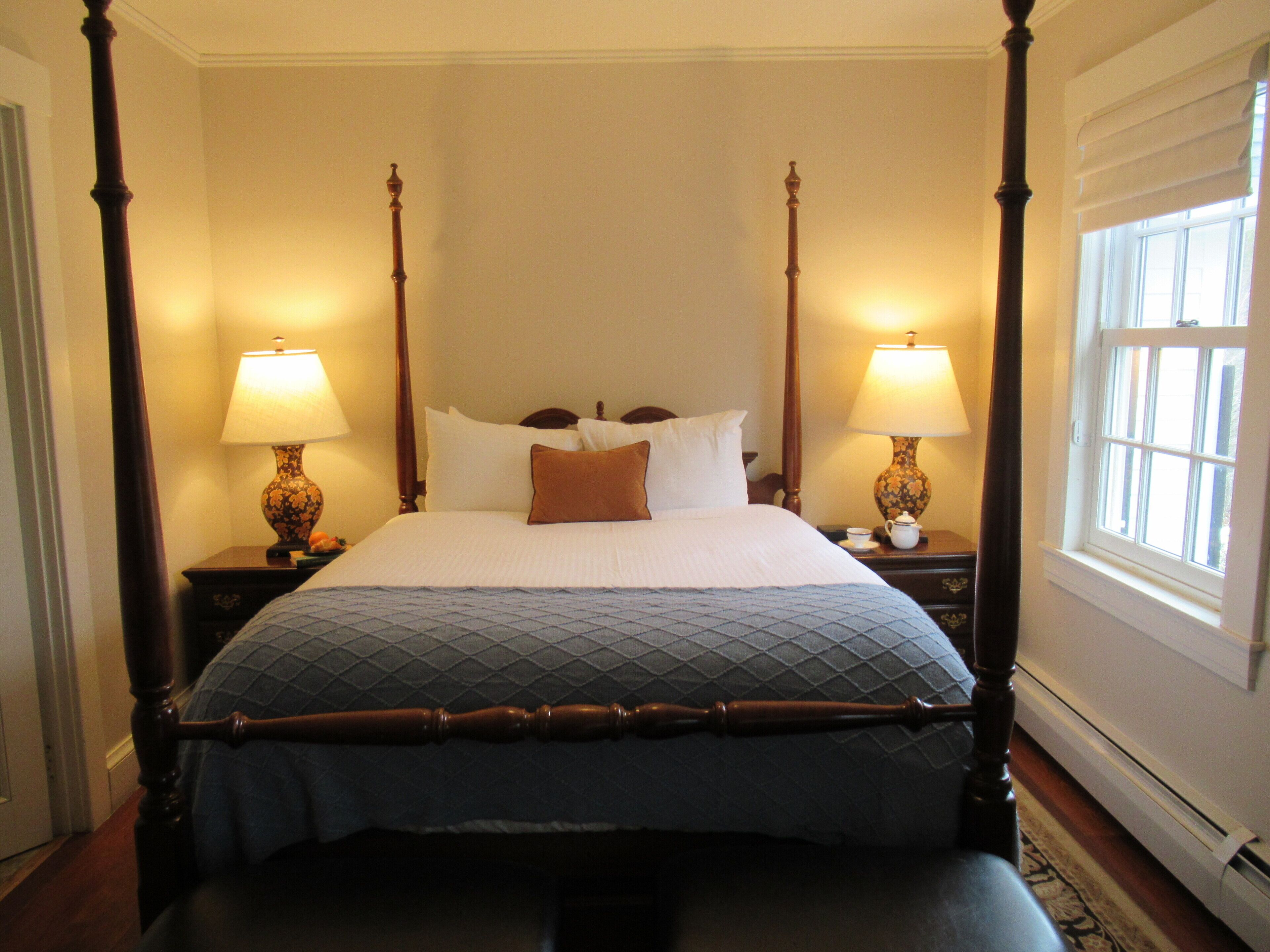A four-poster bed with a quilted bedspread, two bedside lamps, and a window with blinds.