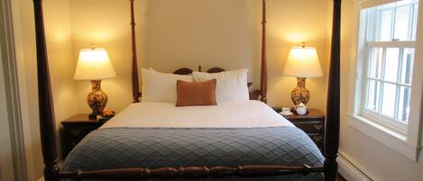 A four-poster bed with a quilted bedspread, two bedside lamps, and a window with blinds.