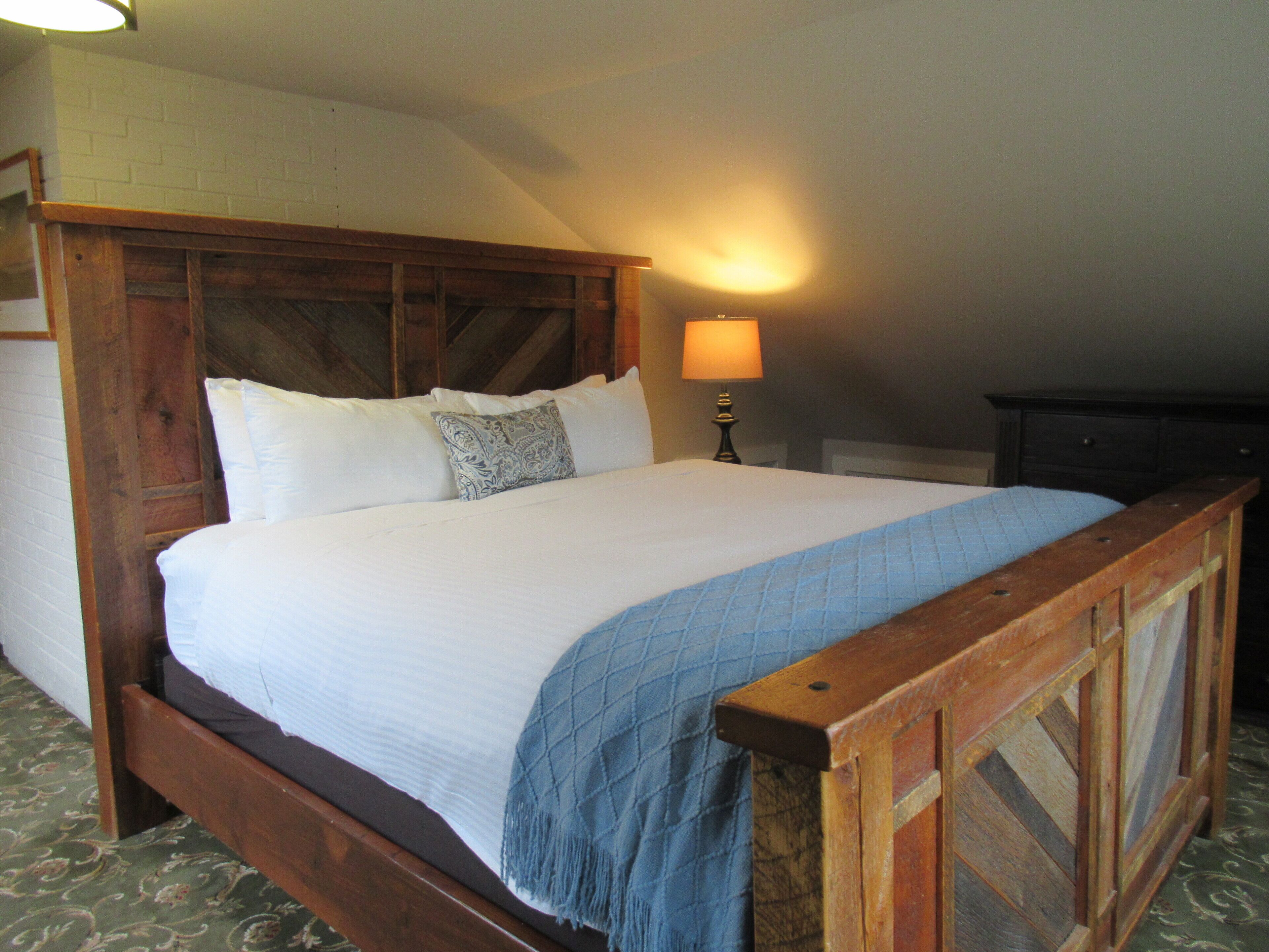 Un lit en bois avec une literie blanche et un plaid bleu, une table de chevet avec une lampe, et une tête de lit en bois.