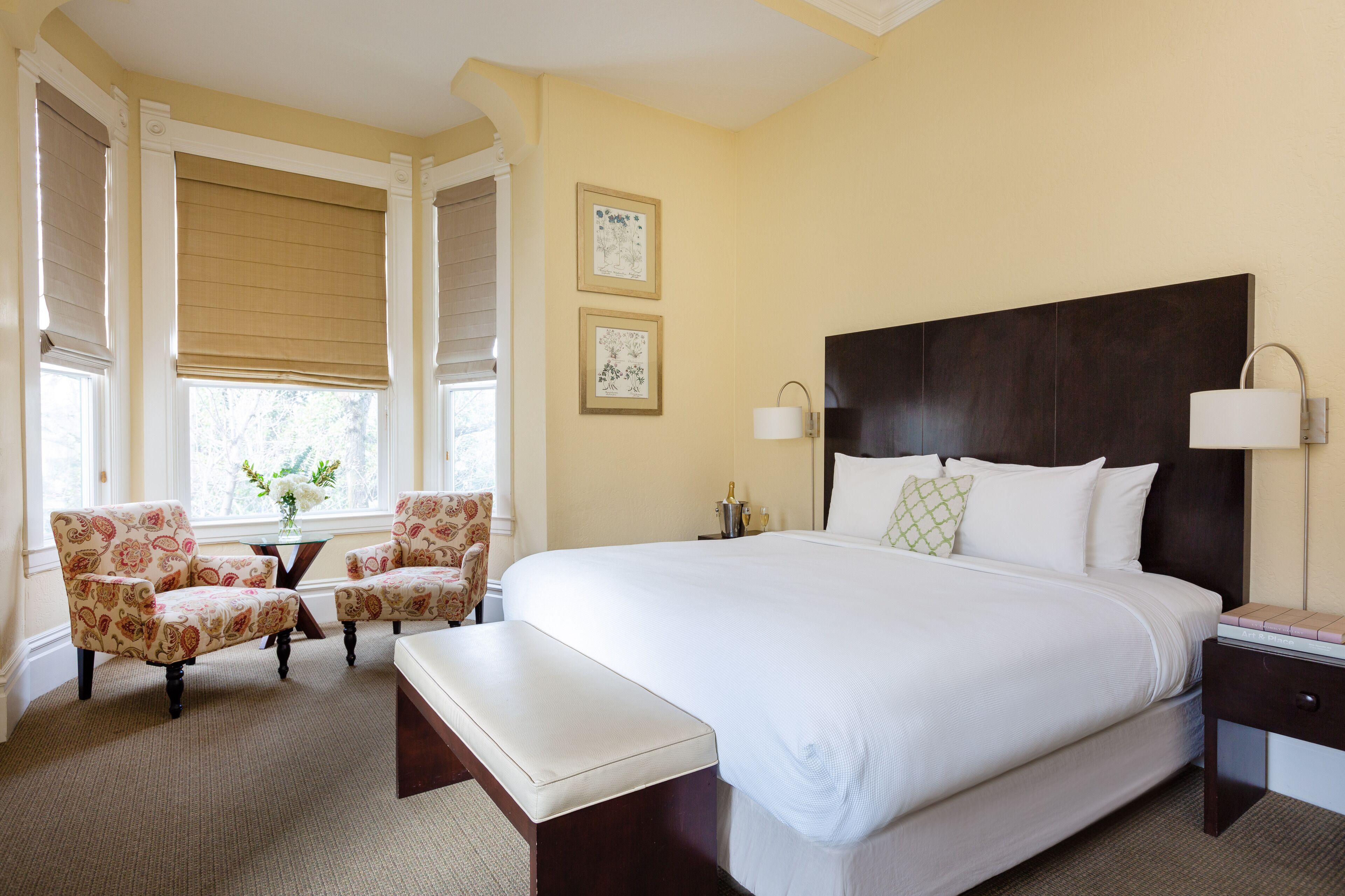Una habitación de hotel con una cama grande, dos sillones, una mesita con un jarrón de flores y una mesita de noche con una lámpara.