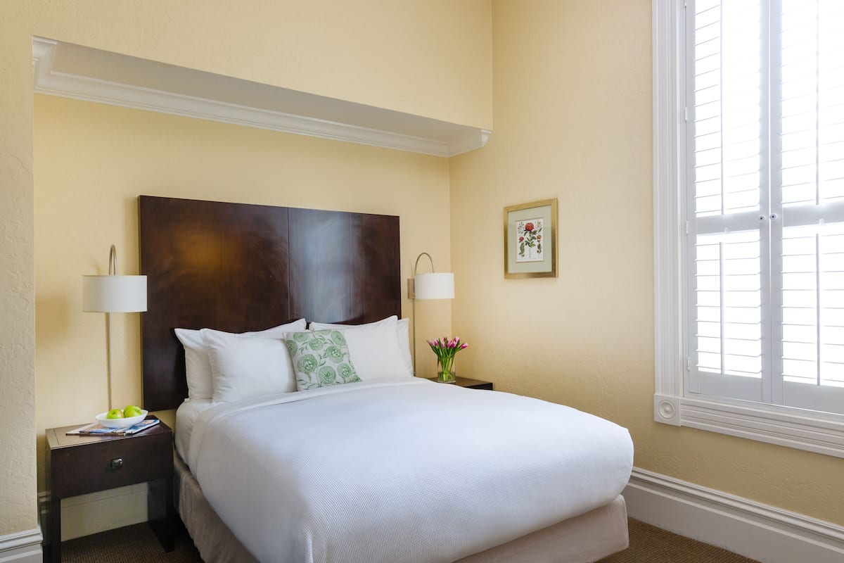 A neatly made bed with white linens, a dark wooden headboard, two bedside lamps, a small table with a bowl of fruit, and a framed picture on the wall.