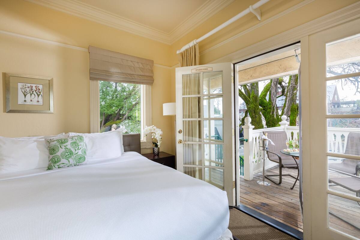 A hotel room with a large bed, a window with a view of greenery, a door leading to an outdoor balcony, and a framed botanical print on the wall.