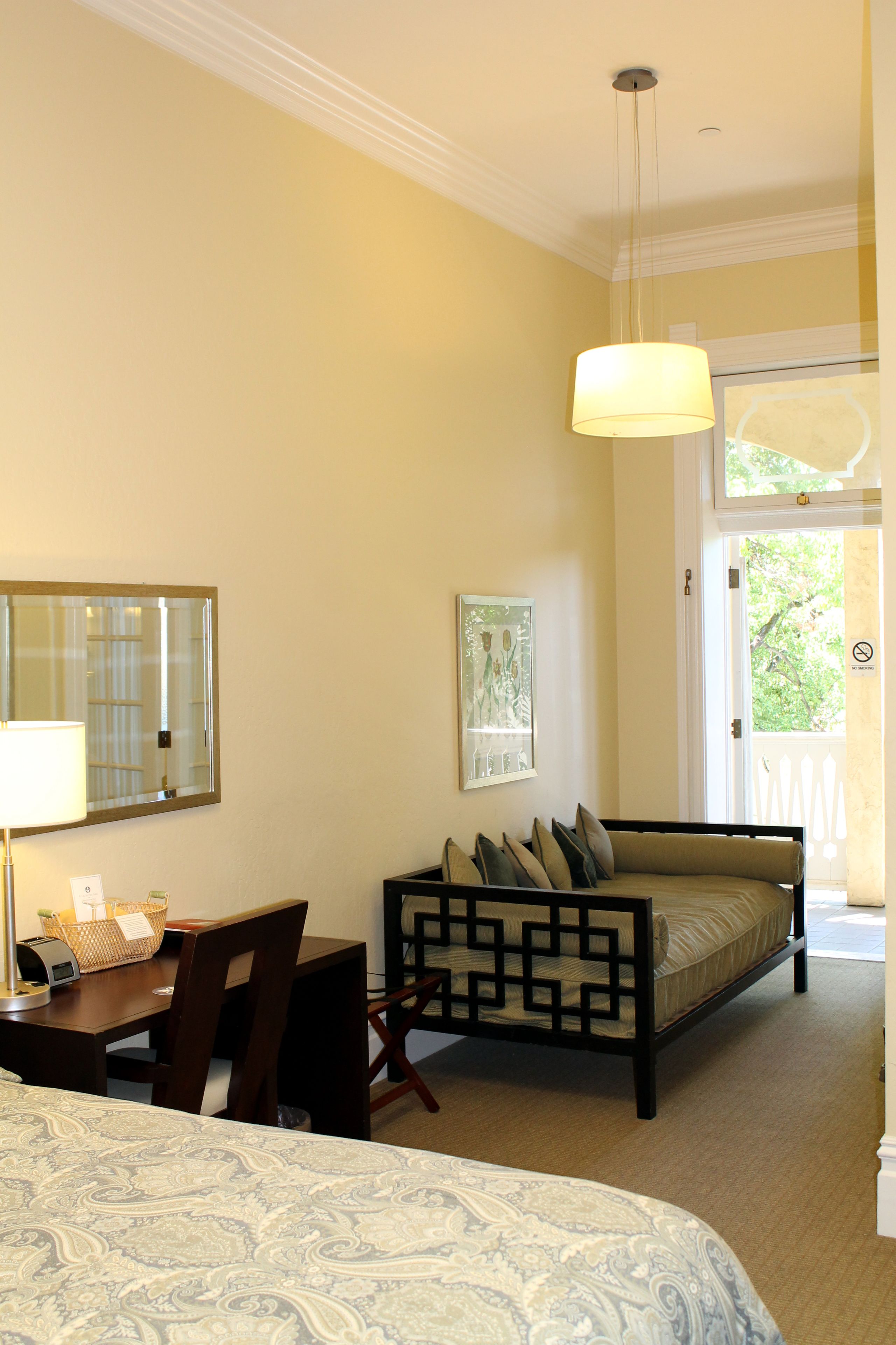 A hotel room with a bed, a desk with a lamp, a chair, a sofa, and a large window.