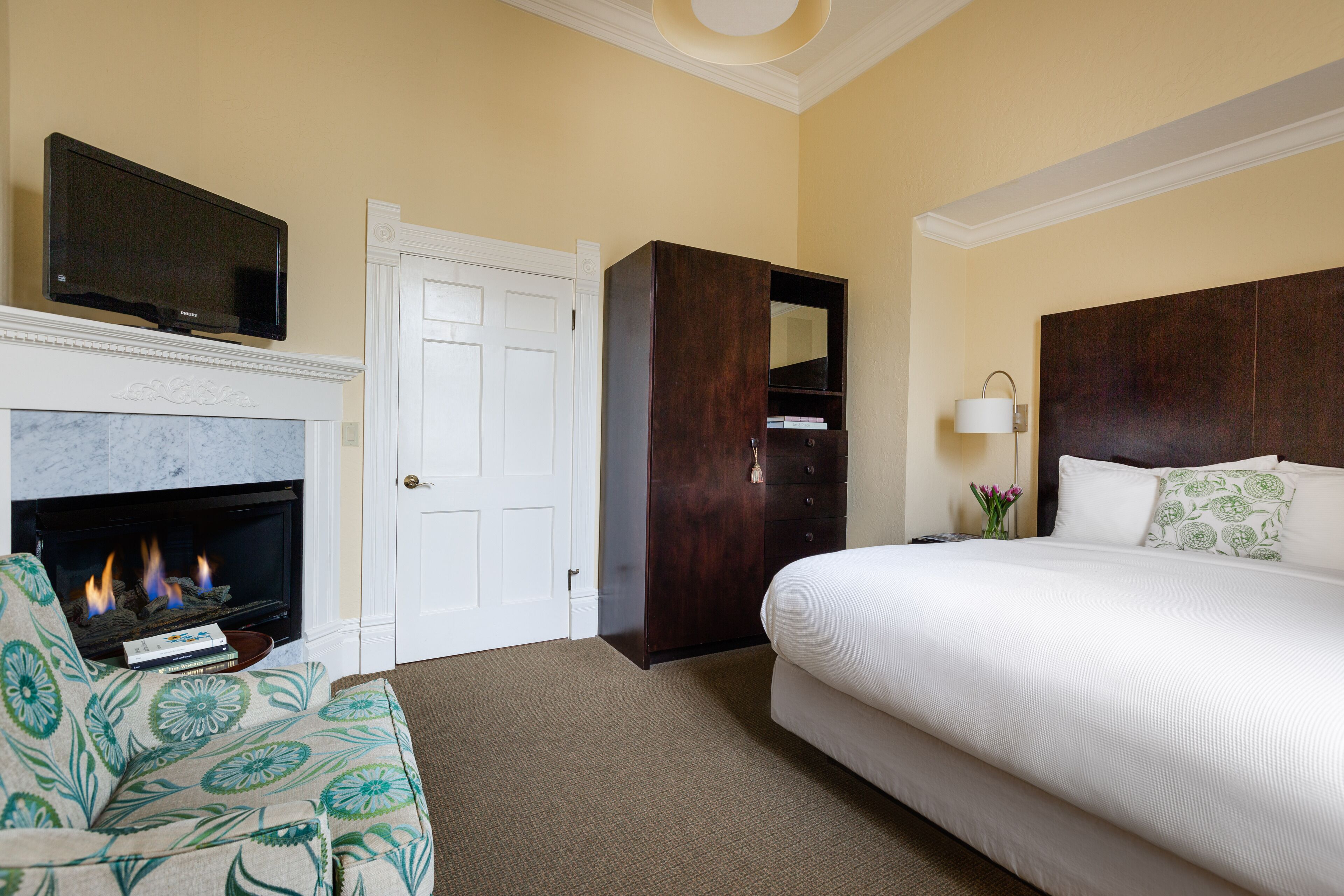 A hotel room with a fireplace, a television mounted above it, a large bed with white bedding, and a dark wooden wardrobe.