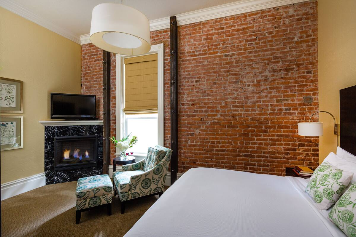 A hotel room with a brick wall, a fireplace, a television, a bed, and a chair.