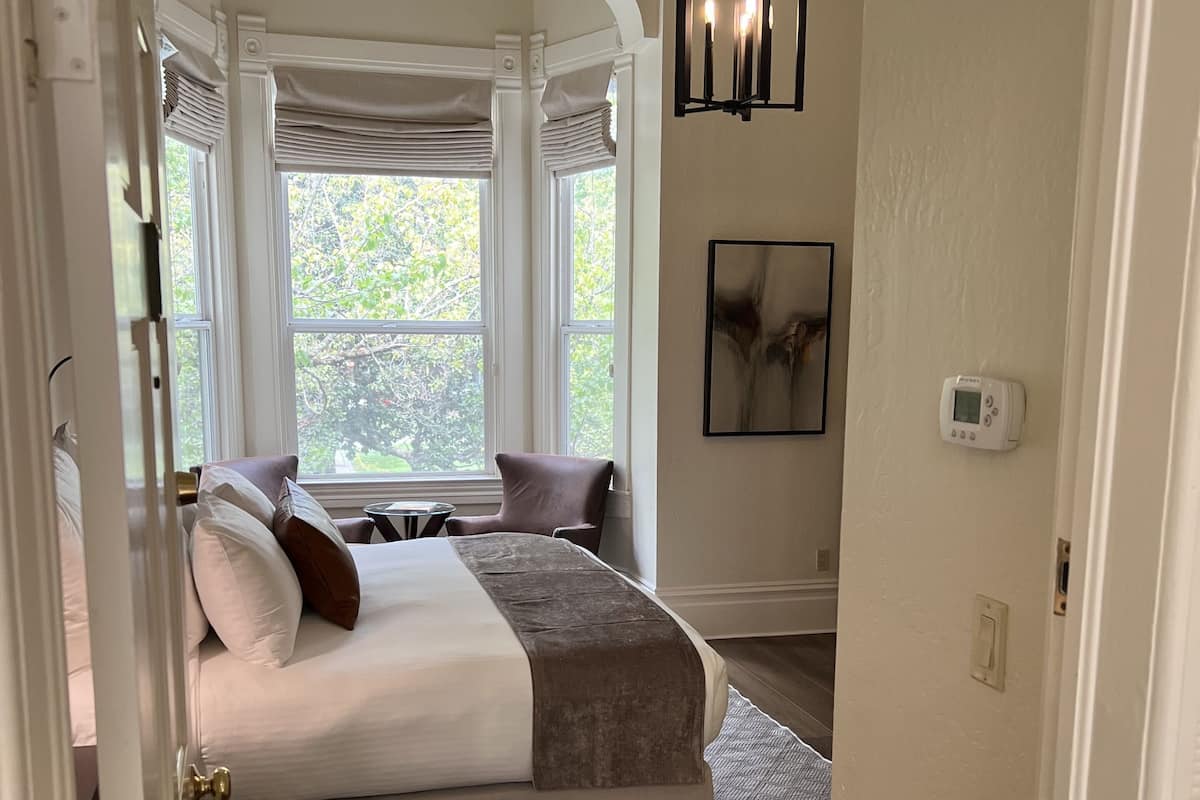 A bedroom with a bed, a window with curtains, a bedside table, and a wall-mounted thermostat.