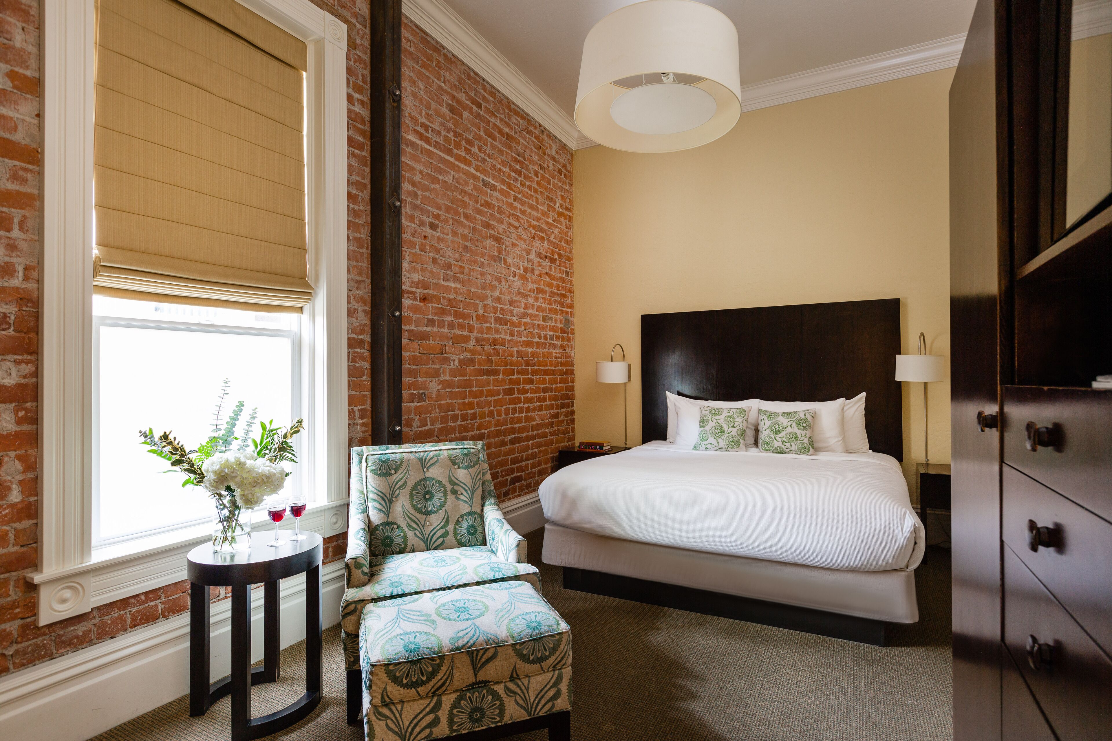 Une chambre d’hôtel avec un mur en briques, un grand lit, une table de chevet, une commode et une fenêtre avec des stores.