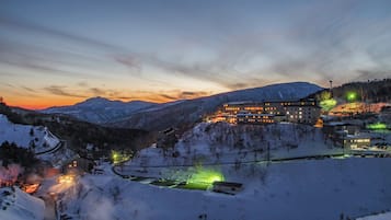 A snow-covered mountain landscape with a large building featuring multiple illuminated windows, surrounded by snow-covered trees and a winding road.