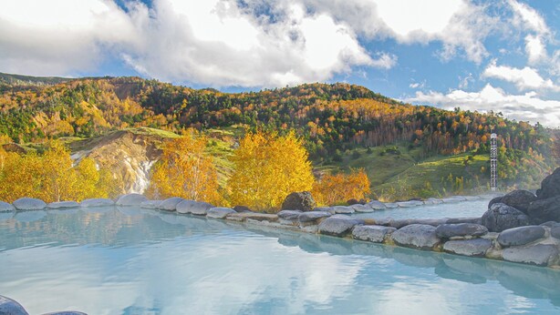 A clear view of a swimming pool with a stone border, surrounded by autumn-colored trees and a mountain in the background. - Manza Prince Hotel (Tsumagoi)