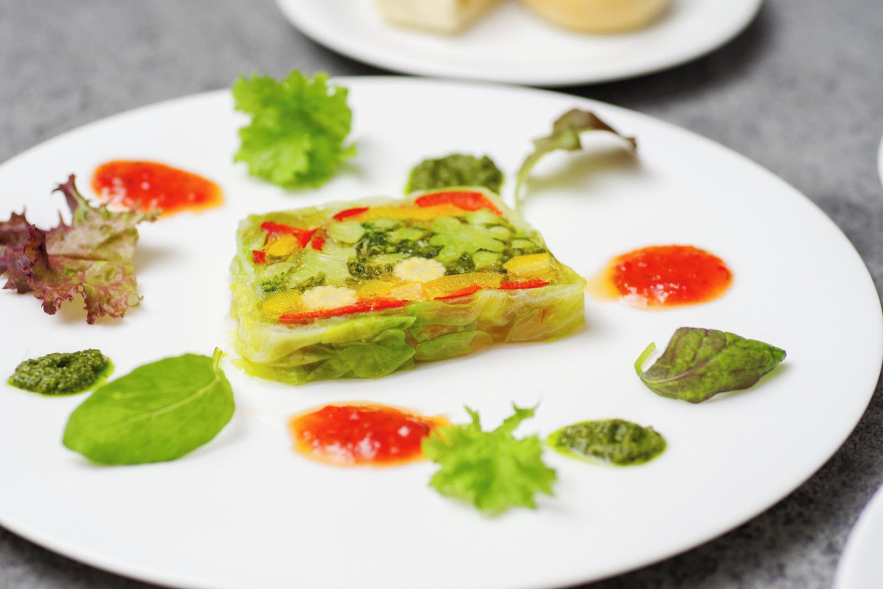 Un plato blanco con un guiso de verduras en capas, adornado con hierbas y salsa roja.
