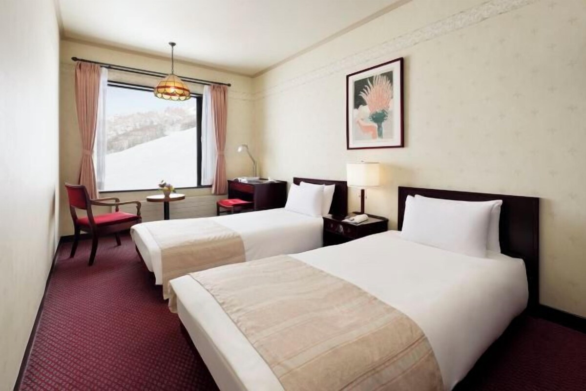 Une chambre d’hôtel avec deux lits, un bureau, une chaise, une lampe et une fenêtre avec des rideaux.