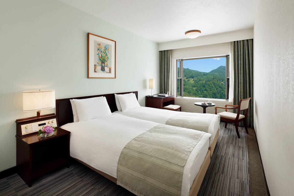 Quarto de hotel com duas camas, mesa de cabeceira com abajur, pequena mesa, cadeira e janela com vista para o verde.