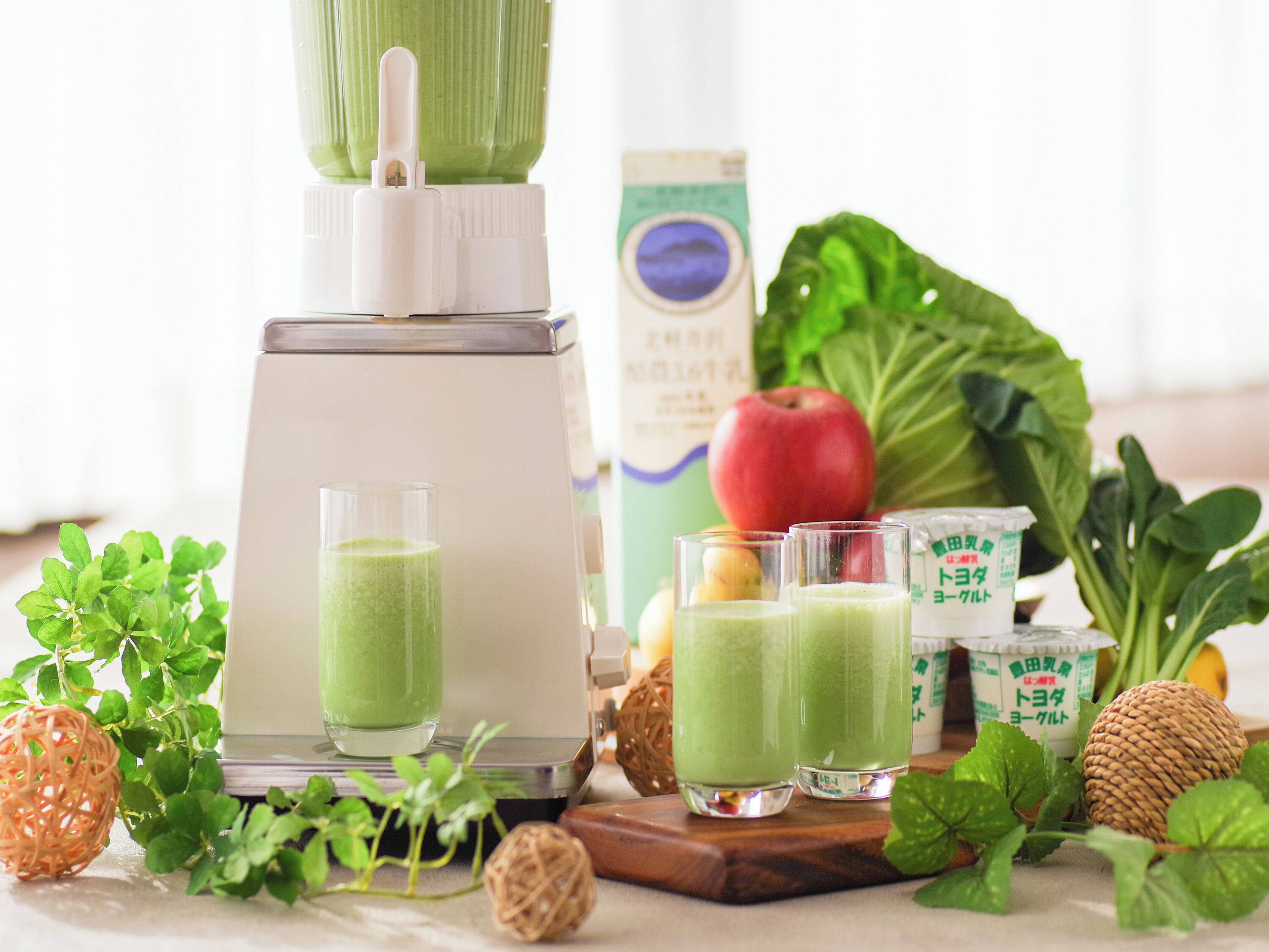 A blender with green smoothie, fresh fruits, and a bottle of milk on a table.