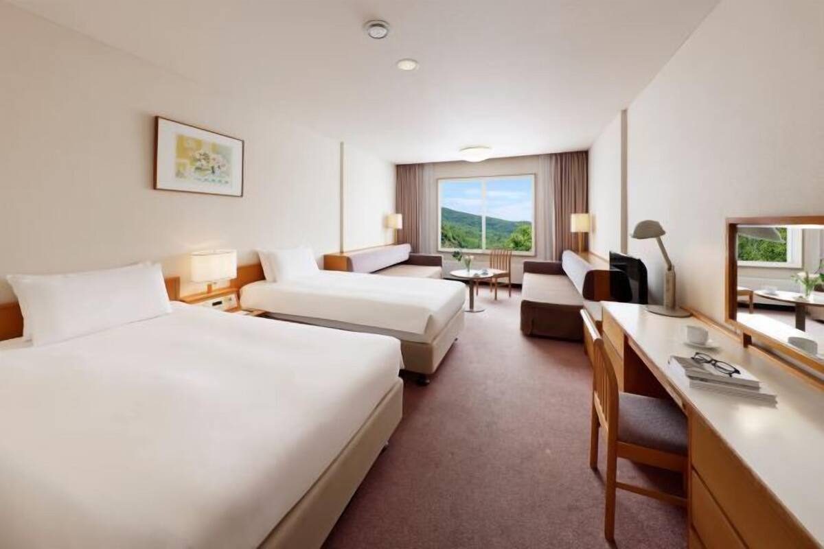 Quarto de hotel com duas camas, uma escrivaninha, uma cadeira e uma janela grande com vista para o verde.