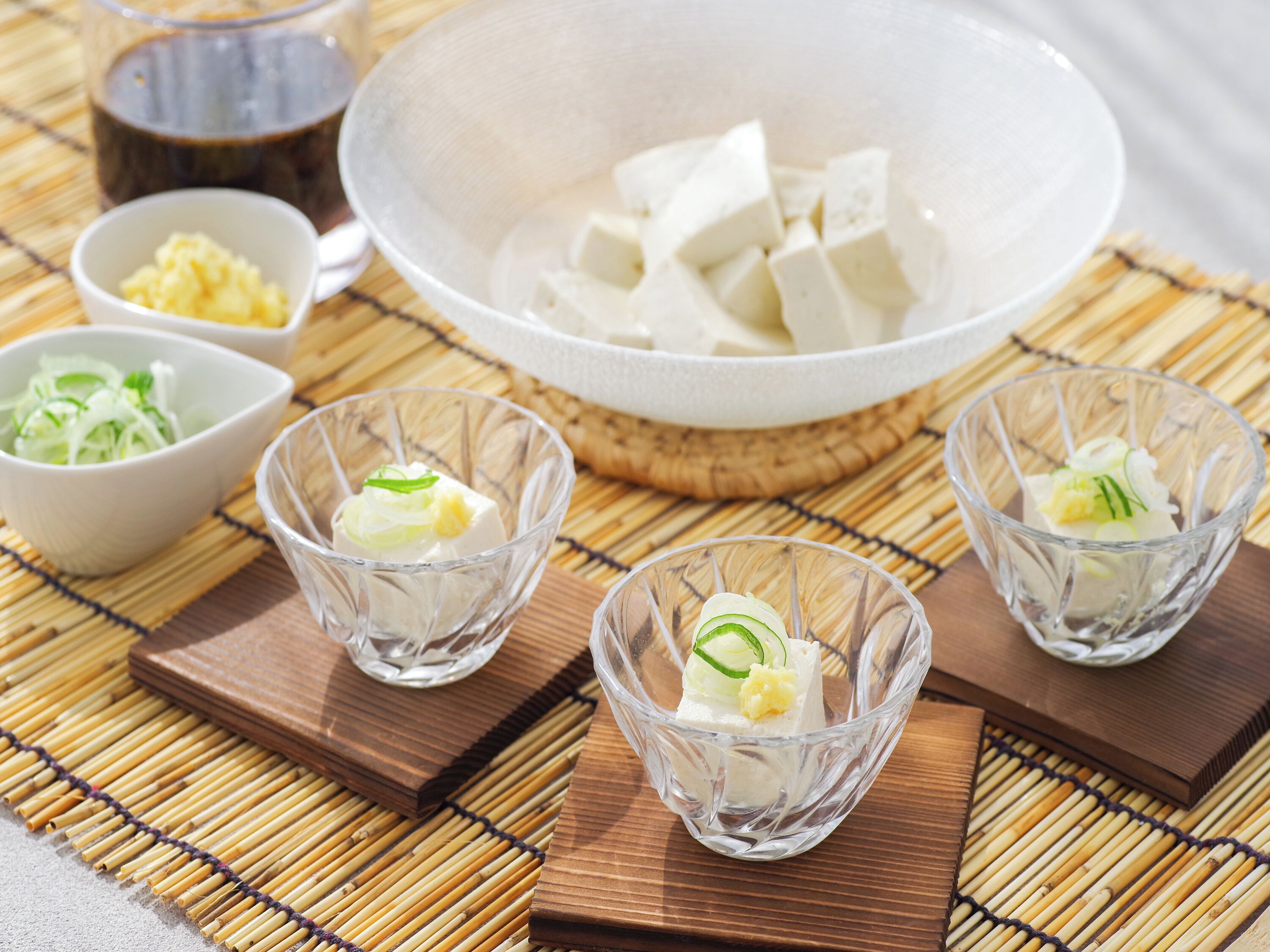Een tafel met een kom tofu, drie kleine glaasjes met tofu en garnering, en een kom gesneden groenten.
