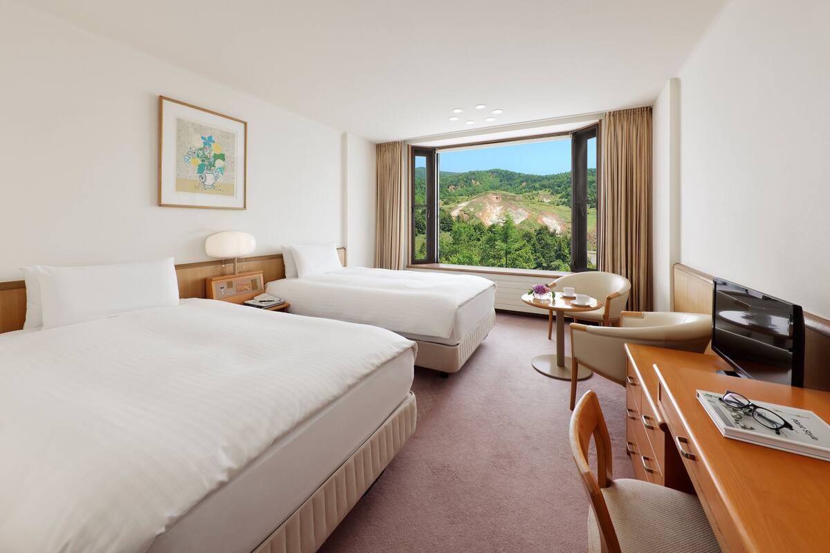 Quarto de hotel com duas camas, uma escrivaninha com cadeira, televisão, janela com vista para o verde e um quadro na parede.