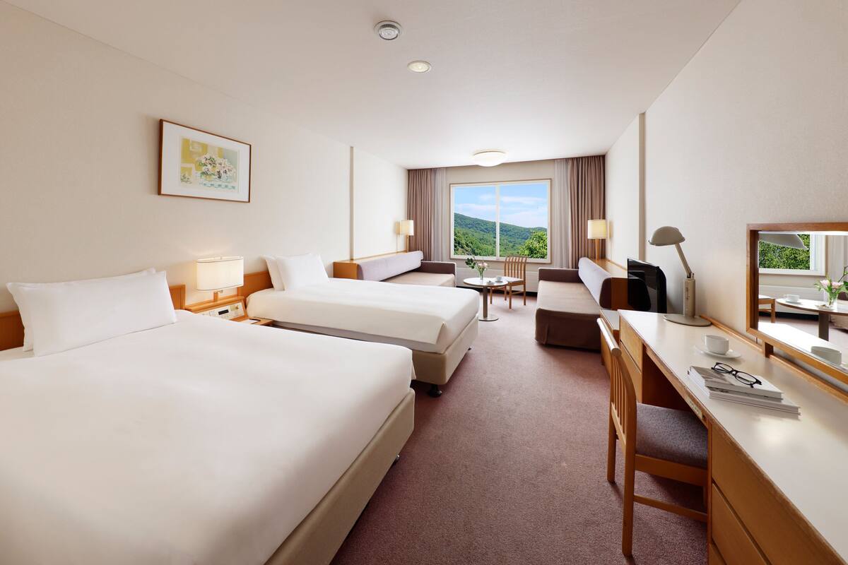 Quarto de hotel com duas camas, uma escrivaninha, uma cadeira, uma mesinha e uma janela com vista para o exterior.