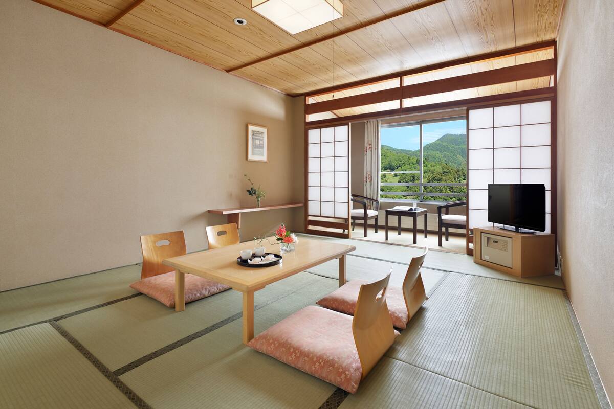 Quarto japonês tradicional com tatames, uma mesa baixa com almofadas, televisão e vista para uma paisagem montanhosa.